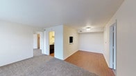 Located at 6495 Piping Rock Ln in Indianapolis, Indiana, this single-floor residence offers a straightforward and functional layout designed for comfortable everyday living. The main living area features a mix of plush carpeting and smooth hardwood-style flooring, creating a natural visual distinction between the relaxation and activity zones of the space. Neutral white walls and standard flush-mount ceiling lighting keep the interior feeling clean and bright throughout. A partial dividing wall separates the living area from the kitchen, which is visible through an open passageway and is equipped with cabinetry and appliances, including a dark-finish dishwasher or range. Overhead ventilation registers are positioned throughout, indicating a central HVAC system for climate control. A hallway extends from the main living space, providing access to additional rooms and what appears to be closet or storage space beyond. The overall design is practical and move-in ready, with a neutral color palette that offers flexibility for personalization. The single-level layout ensures easy accessibility and seamless flow between all areas of the home.