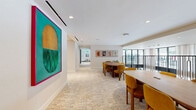 Located at 1430 Lincoln Blvd in Santa Monica, California, this single-floor communal space offers a thoughtfully designed environment that blends functionality with contemporary style. The layout encompasses a spacious open floor plan spread across a generous footprint, seamlessly connecting a living lounge area with a multi-use dining and meeting space.

The entrance lobby features large, floor-to-ceiling black-framed glass walls that flood the interior with natural light while framing views of mature palm trees and a landscaped courtyard beyond. Soft, textured carpeting in neutral tones runs throughout, contributing to a warm and welcoming ambiance. Curved white lounge seating and a petite bistro table create an inviting reception area accented by tall potted plants.

The dining and meeting area is anchored by multiple round wooden tables paired with mustard-yellow upholstered chairs, offering versatile seating configurations. A decorative black metal railing with arch detailing borders an open mezzanine edge, adding architectural character to the space. Two built-in wooden booth alcoves with stone-tiled backdrops and tulip-style side tables provide intimate seating options for smaller gatherings. Framed artwork lines the walls, lending a curated, gallery-like quality. Recessed lighting throughout ensures a bright, even illumination that enhances the space's clean, modern aesthetic.
