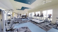This fitness center offers a well-appointed, single-floor layout designed to accommodate a full range of workout routines. The space is bright and airy, with large windows lining the walls and allowing abundant natural light to fill the room, creating an energizing environment for exercise. The ceiling features a distinctive tray design with teal accent panels and recessed lighting, adding a modern, polished aesthetic to the space. Ceiling fans are installed throughout to ensure comfortable airflow. The flooring combines blue exercise mats with patterned carpet tiles, clearly delineating workout zones while adding a sleek, contemporary look. Along one wall, floor-to-ceiling mirrors expand the visual depth of the space and allow users to monitor their form. The cardio zone is well-stocked with multiple treadmills, each equipped with personal display screens, as well as elliptical machines. The strength and free weight area features a full dumbbell rack with a wide range of weights, along with cable machines and additional resistance training equipment. The overall ambiance is clean, modern, and motivating, making this fitness center a functional and visually appealing space for a comprehensive workout experience.