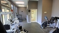 This fitness center is designed to support a well-rounded workout routine within a single-floor layout. The space is finished with durable rubber flooring, ideal for both cardio and strength training activities. Walls are painted in a neutral gray tone, lending the room a modern, focused atmosphere. Large windows along one side of the room allow for generous natural light while offering views of the outdoors, creating an open and energizing environment. The ceiling height adds a sense of spaciousness throughout. Cardio equipment, including treadmills and elliptical machines, lines the windowed wall, while stationary bikes and strength training machines are positioned throughout the remaining floor space, ensuring an efficient and organized use of the area. Overhead fluorescent lighting supplements natural light, keeping the space bright and functional at all hours. An interior doorway connects to an adjacent room, providing additional access within the facility. Informational signage is posted on the walls, offering guidance to users of the space. Overall, this fitness center balances practicality and comfort, making it a well-appointed facility for daily exercise.