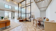 Located at 6850 Foxtail Palm Way in Bradenton, Florida, this single-floor clubhouse space is a masterclass in contemporary design, blending open-concept living with refined architectural details. The centerpiece of the space is a grand lounge area distinguished by dramatically high ceilings, intricate geometric-patterned hardwood flooring, and soaring floor-to-ceiling black-framed glass partitions that allow natural light to cascade freely throughout. Elegant pendant chandeliers and wooden slatted ceiling accents add warmth and texture to the bright, airy atmosphere. Large glass doors open toward views of swaying palm trees, reinforcing the Florida coastal ambiance. A dedicated seating area anchored by a stacked-stone accent wall features a mounted television, a built-in fireplace niche, and rich leather lounge chairs arranged around a marble coffee table. The sleek, galley-style kitchen showcases light wood cabinetry with gold hardware, quartz countertops, open shelving with textured tile backsplash, and statement Edison-style pendant lighting. A casual relaxation nook offers oversized windows with linen drapes, abundant natural light, and a laid-back, inviting feel. Dining and social seating areas are thoughtfully integrated throughout, creating a seamless flow ideal for community gathering and daily leisure.