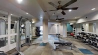 Located at 3101 Rise Dr in Morrisville, NC, this fitness center offers a thoughtfully designed workout environment suited for a wide range of exercise routines. The single-floor facility features an open, airy layout accentuated by multiple ceiling fans that provide steady air circulation throughout the space. Recessed lighting illuminates the room evenly, creating a well-lit environment conducive to focused training sessions. The flooring consists of a patterned combination of neutral and teal-toned tiles, adding a modern aesthetic while offering durability underfoot. Structural columns are integrated into the design, subtly defining different zones within the space. The strength training area is equipped with cable machines, a lat pulldown station, and an adjustable weight bench, catering to a variety of resistance training needs. A mirrored wall along one side enhances the sense of depth while allowing users to monitor their form. On the opposite end, a dedicated functional training area features medicine balls in assorted weights and a stability ball, ideal for dynamic and core-focused workouts. A teal accent wall adds a pop of color, contributing to an energizing atmosphere. Natural light filters in through a window, complementing the interior lighting. Overall, this fitness center provides a comprehensive and inviting space for residents to pursue their health and wellness goals.