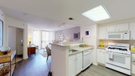 This single-floor residence offers a thoughtfully designed layout that seamlessly connects the kitchen and living areas. The open-concept kitchen features white cabinetry with brushed hardware, light composite countertops, a gas range with overhead microwave, a dishwasher, and a peninsula with an integrated sink that overlooks the living space — ideal for both cooking and entertaining. A skylight panel above the kitchen fills the space with warm overhead illumination.

The living area is bright and welcoming, bathed in natural light from large windows that open to a balcony view. Warm wood-tone flooring, a patterned area rug, and a sophisticated grey accent wall adorned with a cluster of circular mirrors give the space a contemporary, curated character. A dedicated seating and dining zone flows naturally from the living area, enhancing the open feel.

The bedroom is a calm retreat with plush carpeting, a bold blue accent wall, and generous natural light from a large window. It comfortably accommodates a king-sized bed and includes space for a desk or seating area.

The bathroom is clean and functional, finished in white tile with a full shower-tub combination, a white vanity, and ample storage. Consistent neutral tones throughout the residence create a cohesive, modern aesthetic.