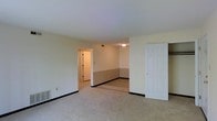 This single-floor residential unit offers a straightforward and functional layout designed for comfortable everyday living. The main living area is finished with wall-to-wall carpeting in a neutral tone, providing a warm and understated foundation throughout the space. Smooth, light-colored walls are complemented by dark baseboards that add a subtle contrast and a clean, defined finish to each room.

A central open passageway connects the main living area to an adjoining room, which features a two-tone wall treatment with a darker wainscoting-style lower section and a lighter upper half, adding a touch of classic detail. This connected room benefits from its own overhead lighting, making it a well-lit and versatile space. A doorway visible beyond leads further into the unit, suggesting additional rooms down a short hallway.

The main living area includes a white six-panel door that opens to a closet, offering practical built-in storage. A floor-level air vent is also present, indicating a centralized heating and cooling system. The overall ambiance is clean, neutral, and move-in ready, with a simple and cohesive aesthetic throughout. The layout flows naturally between spaces, making the unit feel connected and easy to navigate.
