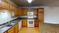 Located at 3374 36th Ave S in Grand Forks, North Dakota, this single-floor unit offers a practical and well-appointed layout designed for comfortable everyday living. A welcoming entry hallway with wood-look flooring leads into an open-concept living and kitchen area. The kitchen is a standout feature, showcasing warm oak cabinetry with ample upper and lower storage, dark granite-style countertops, a central island for additional prep space, and white appliances including a range, over-the-range microwave, dishwasher, and refrigerator. The adjoining living area is finished with plush carpeting and includes a ceiling fan overhead, creating a relaxed and functional common space. The bathroom is well-equipped, featuring oak vanity cabinetry, a granite-style countertop, a large mirror with overhead lighting, and a full tub-shower combination with tile flooring. The bedroom offers carpeted floors and a generous closet with built-in shelving and hanging rods. A dedicated in-unit laundry area includes a full-size washer and dryer with overhead oak cabinetry for additional storage. Throughout the unit, consistent oak woodwork and neutral tones create a cohesive and understated aesthetic.