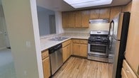 This single-floor residence offers a practical and comfortable living environment designed with everyday functionality in mind. The kitchen is a standout feature, showcasing warm honey-toned wood cabinetry that extends to the upper and lower walls, providing ample storage space. Light stone-patterned countertops run along an L-shaped layout, offering generous prep space and a double-basin stainless steel sink. The kitchen is equipped with modern stainless steel appliances, including a gas range with oven and a dishwasher, complemented by a built-in range hood overhead. The wood-look flooring throughout the kitchen adds a warm, cohesive aesthetic that ties the space together. A peninsula-style counter extends toward the adjacent living area, creating a natural transition between spaces and offering additional utility. Neutral wall tones keep the interior feeling open and clean, while recessed lighting contributes to a well-lit environment. The overall layout is thoughtfully arranged to maximize efficiency, making this a highly functional space suited for comfortable day-to-day living.