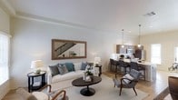 This well-designed two-story residence offers a seamless blend of modern aesthetics and functional living. The main floor showcases an open-concept layout where the living area flows effortlessly into the kitchen and dining space. The living room is bright and inviting, featuring light-toned walls, smooth ceilings, and warm hardwood-style flooring that anchors the space. A large rectangular mirror with a natural wood frame adorns one wall, adding depth and light to the room. The seating area is centered around a circular dark-toned coffee table, complemented by a plush white sofa accented with teal and grey throw pillows, and mid-century modern accent chairs in warm wood tones. The kitchen is equipped with contemporary cabinetry in a warm taupe finish, a light countertop breakfast bar with seating for multiple guests, and pendant lighting overhead that adds a refined touch. Stainless steel appliances and recessed lighting complete the kitchen's polished look. A staircase with sleek metal balusters leads to the second floor, visible in the background and reflected in the mirror, hinting at additional living space above. Natural light filters throughout, enhancing the home's clean, contemporary ambiance.