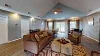 This well-appointed single-floor space showcases a thoughtful open layout with distinct functional zones flowing seamlessly into one another. The centerpiece is a dramatic great room with soaring vaulted ceilings that climb to an impressive peak, fitted with ceiling fans and recessed lighting that create a bright, airy atmosphere. Rich hardwood-style flooring runs throughout, providing a warm, cohesive foundation across all areas.

The main living area is expansive and versatile, featuring a comfortable seating arrangement anchored by a decorative area rug, complemented by a dedicated workspace with built-in shelving. Elegant French doors with large transom windows above flood the space with natural light while providing access to an outdoor area.

A cozy sitting room offers an intimate retreat, furnished with patterned accent chairs arranged around a round central table, with natural light streaming in through window treatments framed by decorative curtains. A separate dining area accommodates a classic round table with leather seating, lit by a contemporary track light fixture.

Throughout the space, soft blue-grey walls create a calm, neutral backdrop that complements the warm furnishings. The combination of vaulted architectural detailing, abundant natural light, and well-defined yet open living zones makes this a comfortable and functional space.