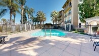 Located at 3250 Market St in Riverside, California, this outdoor amenity space showcases a resort-style pool area defined by its Mediterranean-inspired architectural character. The generously sized swimming pool serves as the centerpiece, its clear turquoise water reflecting the abundant Southern California sunshine. A broad, tiled pool deck surrounds the water, providing ample space for lounging and recreation.

Tall, mature palm trees are planted throughout the area, creating a tropical ambiance and offering natural shade across the deck. The space is framed by a multi-story building featuring warm stucco facades, wrought iron balconies, and terracotta-accented rooflines that reinforce the Mediterranean aesthetic. A charming arched entryway constructed of whitewashed plaster provides a grand passage into the pool courtyard, while a stone-clad pool house with arched openings adds further architectural interest.

A covered pergola structure anchors one end of the space, offering a shaded outdoor retreat ideal for relaxing outside the water. Decorative iron fencing with columned accents encloses the perimeter, lending the area a sense of privacy and elegance. Manicured landscaping with potted plants and garden beds adds color and texture throughout. The overall atmosphere is sunny, inviting, and reminiscent of a private resort retreat in the heart of Riverside.