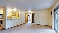 This single-floor space features an open layout connecting the living area to the kitchen and additional rooms beyond. The living area is finished with neutral wall-to-wall carpeting and white walls, creating a clean, understated backdrop. A textured ceiling with flush-mounted lighting provides even illumination throughout the space.

A partial wall with a dark countertop pass-through separates the kitchen from the main living area, allowing visibility into the kitchen without fully opening the floor plan. The kitchen includes a white refrigerator and upper cabinetry finished in a warm wood tone, offering practical storage.

Beyond the living area, a hallway leads to additional rooms, with dark-stained wood doors visible along the corridor. A window at the end of the hallway introduces natural light into the interior. Dark wood baseboards provide contrast against the light-colored walls and carpeting throughout.

The overall ambiance is practical and neutral, with a simple, functional design. The open sightlines between the living area, kitchen, and hallway contribute to an efficient use of space, making the layout feel connected and easy to navigate.