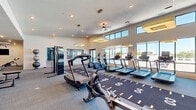 This expansive fitness center is designed to deliver a comprehensive, high-energy workout experience within a thoughtfully planned open-floor layout. The space is defined by soaring ceilings with a clerestory window system that wraps the upper perimeter, flooding the interior with natural light and offering open outdoor views. Large floor-to-ceiling windows along one wall further enhance the bright, airy atmosphere.

The cardio zone features a row of treadmills, ellipticals, and stationary bikes, all positioned along an indoor track runner mat marked with distance indicators — a standout architectural and functional detail. Stylish square pendant light fixtures and ceiling fans are suspended overhead, complementing the recessed lighting throughout.

The strength training area is equally well-equipped, housing multi-station cable machines, a free weights section with a full dumbbell rack, a Smith machine with bench, and functional training accessories including battle ropes and plyo boxes. A vibrant mural adds bold color to the brick-accented accent wall, lending the space a motivating, contemporary energy.

A dedicated room with teal accent walls, a wall-mounted TV, and a mirrored wall provides a focused area for targeted strength work. Soft carpet tile flooring spans the entire facility, offering comfort and durability throughout.