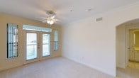 This single-floor space offers a clean, well-maintained living area highlighted by several thoughtful architectural details. The main living space is finished with plush, neutral-toned carpeting that adds warmth underfoot, while crisp white crown molding runs along the perimeter of the ceiling, lending a polished, classic touch to the interior.

A ceiling fan with an integrated light fixture is centrally mounted, providing both air circulation and ambient lighting. The walls are painted in a soft, warm neutral tone, creating a calm and inviting atmosphere that suits a variety of interior styles.

Natural light is a standout feature of this space, delivered through a set of glass-paneled French doors that lead out to a balcony. A narrow sidelight window beside the doors further enhances the brightness of the room. The doors offer views of the surrounding outdoor environment, adding a sense of openness and connection to the exterior.

Practical elements include a ceiling-mounted HVAC vent for climate control and a standard electrical outlet along the baseboard. The space is unfurnished, making it easy to envision a wide range of furniture arrangements and personalization options.