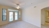 This single-floor space offers a clean, well-maintained living area highlighted by several thoughtful architectural details. The main living space is finished with plush, neutral-toned carpeting that adds warmth underfoot, while crisp white crown molding runs along the perimeter of the ceiling, lending a polished, classic touch to the interior.

A ceiling fan with an integrated light fixture is centrally mounted, providing both air circulation and ambient lighting. The walls are painted in a soft, warm neutral tone, creating a calm and inviting atmosphere that suits a variety of interior styles.

Natural light is a standout feature of this space, delivered through a set of glass-paneled French doors that lead out to a balcony. A narrow sidelight window beside the doors further enhances the brightness of the room. The doors offer views of the surrounding outdoor environment, adding a sense of openness and connection to the exterior.

Practical elements include a ceiling-mounted HVAC vent for climate control and a standard electrical outlet along the baseboard. The space is unfurnished, making it easy to envision a wide range of furniture arrangements and personalization options.
