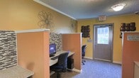 This single-floor space offers a practical and inviting environment designed to serve visitors and residents alike. The interior is finished with warm yellow and terracotta-toned walls, accented by decorative wire floral wall art and a mosaic tile detail that adds texture and visual interest to the workstation divider. The built-in computer workstation area features a granite-style countertop, desktop computer, keyboard, and ergonomic seating, providing a dedicated space for productivity or information access. A secondary chair station is visible in the background, offering additional workspace capacity. The flooring is covered in a durable, neutral-toned carpet that complements the overall color scheme. Crown molding runs along the ceiling line, lending a polished, finished look to the room, while a textured ceiling adds depth overhead. Natural light filters in through a windowed entry door, brightening the space and connecting it to the outdoors. A clearly marked exit sign and security surveillance notice reflect a commitment to safety and transparency. Overall, the space strikes a balance between functional utility and approachable, cheerful design.