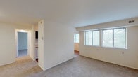 This single-floor residence offers a straightforward, functional layout organized around a central living area. The main living space is generously sized, finished with neutral wall-to-wall carpeting and painted in crisp white tones. A partial dividing wall separates the living zone from an adjoining area, adding subtle architectural definition to the open floor plan. Multiple windows fitted with horizontal blinds allow natural light to filter in, providing pleasant views of surrounding greenery.

The galley-style kitchen runs along a corridor layout, featuring dark espresso-toned cabinetry on both sides, white countertops with an integrated sink, and a full-size refrigerator. Wood-look flooring runs throughout the kitchen, and a window at the far end adds natural light to the workspace.

Two bedrooms are carpeted and finished in the same neutral palette. Each includes a closet—one is a standard reach-in with a rod, and the other is a walk-in closet accessible through a dedicated door, providing ample storage.

The bathroom includes a single-sink vanity with a dark cabinet base, a mirror with overhead lighting, and a full tub-shower combination with white tile surround. Tile flooring completes the clean, functional design of the space.