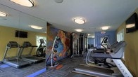 This single-floor fitness center offers a comprehensive workout environment designed to accommodate a variety of exercise routines. The space is anchored by a striking, large-scale colorful mural depicting a dynamic athletic figure, serving as a bold focal point that energizes the room. Full-length mirrors line one wall, visually expanding the space and allowing users to monitor their form during workouts. Ceiling-mounted flush light fixtures provide consistent, even illumination throughout the facility. The cardio section features multiple treadmills and an elliptical trainer, positioned to make use of the available floor space efficiently. A multi-station cable and weight machine occupies the central area, offering a range of strength training options. A dedicated dumbbell and free weights rack is also visible, catering to users of varying fitness levels. The flooring consists of durable, patterned carpet tiles suited for a high-traffic fitness environment. Painted in contrasting shades of yellow and blue, the walls contribute a lively, motivating atmosphere. An emergency exit with clearly marked signage ensures safety compliance. Overall, the space is thoughtfully arranged to provide a functional, inviting, and well-organized fitness experience.