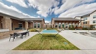 This two-floor residential community is defined by its thoughtfully designed outdoor amenity spaces and cohesive architectural identity. The exterior showcases warm brick facades, standing seam metal roofing, and large-paned windows that allow natural light to flow throughout the buildings. A grand covered entry corridor welcomes residents with vaulted wood ceilings, pendant lighting, and paver stone flooring that sets an inviting tone from arrival.

At the heart of the community lies a resort-style pool area featuring a spacious swimming pool, an in-pool lounge shelf with reclining chaise chairs, and an adjoining spa. A substantial wood-beamed pergola serves as a central outdoor pavilion, providing shade and a social anchor to the pool deck. Surrounding the pool, lounge chairs and seating areas are arranged for relaxed outdoor living.

Beyond the pool, a dedicated hammock lawn area offers a peaceful retreat, flanked by ornamental grasses and landscaped garden beds that enhance the natural ambiance. A fire pit lounge with Adirondack-style seating provides an inviting outdoor gathering spot. Covered terraces with tile flooring, glass railings, and wood plank ceilings extend the communal living experience, offering additional furnished areas for dining and socializing. Lush, low-maintenance native plantings frame the buildings throughout, completing the polished, community-centered outdoor environment.
