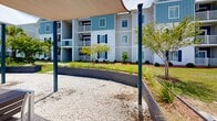Located at 50 Newport Boulevard in Port Wentworth, Georgia, this single-story outdoor amenity space serves as a central hub for community living. The grounds are thoughtfully designed with a resort-inspired aesthetic, blending functional gathering areas with attractive landscaping throughout.

At the heart of the outdoor space is a community pool, enclosed by a sleek black iron fence and complemented by poolside seating and striped umbrellas. Adjacent to the pool, a gravel courtyard hosts a well-appointed pergola structure supported by teal-painted steel posts, providing shaded outdoor dining areas with tables and chairs arranged for social gatherings. Sail-shade canopies add additional sun coverage over select zones.

Raised planter beds constructed from dark composite materials introduce structured greenery, filled with ornamental grasses and shrubs that add visual depth. Palm trees and mature deciduous trees are interspersed throughout, lending a coastal Southern character to the setting.

A white-sided amenity building with a hipped roof and French doors anchors one end of the courtyard, offering direct access to interior facilities. Wide concrete walkways connect all areas and lead to the multi-story residential buildings nearby, which feature blue-and-white coastal-style facades with balconies. The overall ambiance is clean, welcoming, and well-maintained, reflecting a thoughtfully planned community environment.