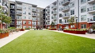 Located at 511 S Mangum St in the heart of Durham, NC, this 3-floor residential community is centered around a beautifully designed interior courtyard that serves as the social and recreational hub of the property. The building's exterior showcases a striking combination of classic red brick and modern light-panel siding, complemented by sleek black-railed balconies that extend from units on every level, offering residents elevated views of the lush courtyard below.

The courtyard itself is a standout feature, anchored by a generous, well-maintained lawn that provides open green space for relaxation and recreation. Surrounding the lawn, a wide paved walkway connects multiple seating areas outfitted with Adirondack chairs, dining sets, and umbrella-shaded tables — creating inviting spaces for gathering or quiet outdoor enjoyment. A built-in corten steel fireplace adds a distinctive architectural accent and a warm focal point for evening use.

Raised planter beds filled with colorful seasonal plantings and ornamental shrubs line the courtyard perimeter, contributing to the vibrant, manicured landscaping throughout. Ground-floor units feature private patios that open directly onto the courtyard, while upper-level units enjoy private balconies. The cohesive design blends urban contemporary aesthetics with comfortable communal living in one of Durham's dynamic neighborhoods.