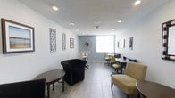 This single-floor business center is designed to provide a functional yet inviting environment for productivity and professional use. The space features a clean, modern aesthetic with light gray walls, a contrasting blue-gray accent wall, and wood-look tile flooring that adds warmth throughout. Recessed and track lighting illuminate the room evenly, complementing the natural light that filters in through a prominent window. On one side, a row of ergonomic task chairs in a distinctive yellow-green hue lines a built-in desk surface, providing multiple dedicated workstations equipped for computer use. On the opposite side, a cozy lounge area features a classic tufted black armchair with nailhead trim, a small round side table, and additional guest seating, creating an informal space for relaxed collaboration or a brief reprieve. A tall bistro table near the accent wall offers yet another casual work or meeting option. Decorative touches, including framed wall art, a starburst mirror, and thoughtfully placed accents, give the space a polished, welcoming character. The layout is open and well-organized, making it easy to navigate between the lounge and work areas. Overall, the business center strikes a balance between efficiency and comfort, catering to a variety of professional needs.