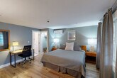Located at 473 Main St in Barnstable, MA, this thoughtfully designed one-bedroom, one-bathroom space offers a comfortable and functional layout with a coastal aesthetic. The bedroom serves as the heart of the unit, featuring warm hardwood flooring, soft blue-gray walls, and a queen bed dressed in crisp, neutral bedding flanked by wooden nightstands with stylish table lamps. A wall-mounted split AC/heating unit ensures year-round climate comfort. Natural light filters in through windows dressed with layered curtains and blinds, providing a bright yet relaxed ambiance. A dedicated workspace with a desk and ergonomic chair adds practicality to the room. The bedroom also includes a walk-in closet with shelving and luggage storage. Along one wall, a media console houses a large flat-screen TV, mini-refrigerator, and microwave, complemented by a white barn-style cabinet offering additional storage and a coffee station. A framed mirror and decorative artwork complete the coastal-inspired décor. The private bathroom features classic white tile flooring with a red diamond accent pattern, a combined tub and shower, a single vanity, and a bright window that fills the space with natural light. This efficiently designed space offers a complete and comfortable retreat in the heart of Cape Cod.