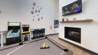 This single-floor amenity space is thoughtfully designed to encourage socializing and relaxation. The game room serves as a focal point, boasting soaring ceilings that amplify the open atmosphere. A pool table anchors the space, complemented by classic arcade machines and high-top seating along a narrow bar-style table. A shiplap-framed fireplace with a wood mantel and wall-mounted television adds warmth, while a distinctive collection of decorative turtle shell wall art lends a unique, artistic character to the room.

Adjacent to the game area, a lounge nook features a bistro-style table with teal nailhead-trim chairs beneath a pendant light, framed by patterned drapes and a natural-light window. Open shelving along one wall provides both decorative and functional storage.

The beverage station rounds out the space, offering a dedicated coffee machine, mini refrigerator, white subway tile backsplash, and organized condiment storage on white countertops with grey cabinetry below. A wall-mounted television completes this functional corner. Light wood-look flooring flows consistently throughout, tying the various areas together cohesively.