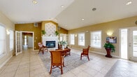 Located in Vero Beach, Florida, this single-story clubhouse and fitness center offers a well-rounded community amenity space designed for comfort and functionality. The welcoming entrance opens into a generously sized lounge area finished with large-format tile flooring, soaring vaulted ceilings with recessed lighting, and abundant natural light streaming through multiple windows. A prominent fireplace with a decorative tile surround serves as the focal point of this central gathering area, which is furnished with rattan-style chairs and a coffee table set atop an area rug, creating an inviting atmosphere.

The dining room features patterned carpet, warm golden-toned walls with white wainscoting and framed wall panels, and elegant bistro-style table and chair sets — ideal for casual community gatherings. Adjacent to it is a secondary dining area with a pass-through kitchenette and bar seating. The office provides a dedicated computer workstation area with built-in cabinetry and multiple workstations for resident use. The exercise room is well-equipped with treadmills, an elliptical, a multi-station strength machine, and a recumbent bike, all beneath ceiling fans for comfort. Multiple connecting hallways tie each area of the facility together seamlessly, offering easy navigation throughout.