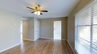 This single-story residence features two bedrooms and one bathroom within a well-utilized floor plan. The main living area is finished with warm-toned hardwood-style flooring that extends throughout, providing a clean and cohesive aesthetic. Neutral gray walls complement the light flooring, creating a bright and versatile backdrop for any décor style. A ceiling fan with an integrated light fixture is centrally mounted, offering both airflow and ambient lighting to the space. White baseboards and door trim provide crisp contrast against the neutral walls, adding a polished finishing touch. HVAC venting is integrated into the ceiling, ensuring efficient climate control. Doorways connect the living area to adjacent rooms, allowing for a natural flow through the home. Windows fitted with blinds invite natural light while maintaining privacy. The overall design is clean and straightforward, with practical finishes that are easy to maintain. This move-in-ready home provides an efficient and comfortable living environment suited for a range of lifestyles.