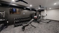 Located at 412 12th Ave SE in Minneapolis, MN, this fitness center is a compact yet comprehensively outfitted space designed to support a full range of workout routines. The facility is divided into two distinct zones, each with its own aesthetic and purpose. The primary workout area features dark, matte-finished walls, textured carpet tile flooring, and exposed ductwork running along the ceiling, giving it an industrial, high-energy atmosphere. Ceiling fans and recessed lighting provide consistent airflow and visibility throughout. This zone is equipped with commercial-grade treadmills featuring digital displays, elliptical machines, a multi-station cable machine, and a full dumbbell rack spanning a wide weight range, complemented by an adjustable weight bench. A wall-mounted flat-screen TV and mirrored wall panels add functionality and visual depth to the space. A secondary, brighter area features light hardwood flooring, a drop ceiling with recessed lighting, and large glass doors that invite natural light. This section houses a power rack with a barbell and weight plates, a cable crossover machine, TRX suspension trainers, resistance bands, and additional free weight accessories, making it ideal for strength and functional training. Together, the two areas create a versatile and well-appointed fitness environment.