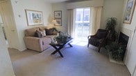 This single-floor residence offers a comfortable and well-appointed living space designed with a warm, neutral aesthetic throughout. The living room serves as the centerpiece of the home, featuring plush wall-to-wall carpeting that adds a sense of coziness underfoot. Large sliding glass doors flood the room with abundant natural light and provide direct access to a private balcony, offering pleasant outdoor views. The space is thoughtfully arranged with generous seating, including a cushioned sofa accented with decorative pillows and a dark wicker armchair, creating an inviting atmosphere for relaxation or entertaining. A stylish X-base coffee table sits at the heart of the seating area, topped with tasteful decorative accents. Soft, neutral wall tones complement the light-colored carpeting, while framed artwork adds a refined touch to the walls. A classic table lamp provides warm ambient lighting, enhancing the overall sense of comfort. Sheer curtains and a structured valance frame the sliding doors elegantly, balancing privacy with the room's bright and airy feel. The overall ambiance is calm, approachable, and move-in ready, making it an ideal space for those seeking a tranquil and well-maintained home environment.