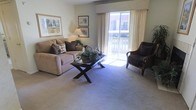 This single-floor residence offers a comfortable and well-appointed living space designed with a warm, neutral aesthetic throughout. The living room serves as the centerpiece of the home, featuring plush wall-to-wall carpeting that adds a sense of coziness underfoot. Large sliding glass doors flood the room with abundant natural light and provide direct access to a private balcony, offering pleasant outdoor views. The space is thoughtfully arranged with generous seating, including a cushioned sofa accented with decorative pillows and a dark wicker armchair, creating an inviting atmosphere for relaxation or entertaining. A stylish X-base coffee table sits at the heart of the seating area, topped with tasteful decorative accents. Soft, neutral wall tones complement the light-colored carpeting, while framed artwork adds a refined touch to the walls. A classic table lamp provides warm ambient lighting, enhancing the overall sense of comfort. Sheer curtains and a structured valance frame the sliding doors elegantly, balancing privacy with the room's bright and airy feel. The overall ambiance is calm, approachable, and move-in ready, making it an ideal space for those seeking a tranquil and well-maintained home environment.