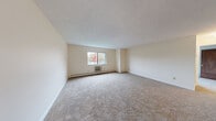 Located at 17 Conant Street in Beverly, Massachusetts, this apartment offers a practical and comfortable layout across a single floor, with two bedrooms, one bathroom, and a dedicated kitchen. The expansive living area is finished with neutral-toned wall-to-wall carpeting, creating a warm and inviting atmosphere. Large windows allow natural light to fill the room while framing views of the surrounding neighborhood and seasonal foliage. Baseboard heating and a wall-mounted A/C unit ensure year-round comfort. The generously sized primary bedroom features two wide windows on adjacent walls, maximizing light and outdoor views, and is complemented by a large walk-in closet with built-in shelving and hanging rods. The kitchen is equipped with rich wood cabinetry, white laminate countertops, a tile backsplash, hardwood flooring, and white appliances including a dishwasher and electric range. A peninsula counter adds additional prep space. The full bathroom features mosaic tile flooring, a white vanity, and a walk-in shower with accessibility grab bars. An additional closet provides extra storage. The layout flows efficiently, making smart use of the available space throughout.