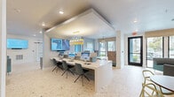 Located in Longwood, Florida, this single-floor leasing and amenity center offers a well-appointed, contemporary environment designed to welcome and inform visitors. The space features terrazzo flooring throughout, lending a polished, cohesive aesthetic that ties together its distinct functional zones.

At the heart of the space is a prominent workstation counter lined with multiple computer terminals and wall-mounted displays, illuminated by a striking geometric pendant chandelier. Adjacent to this area, a comfortable lounge features teal upholstered seating, colorful ottomans, and vibrant artwork, creating an inviting atmosphere for guests.

A dedicated coffee bar area is outfitted with grey shaker-style cabinetry, an eye-catching gold and white patterned tile backsplash, a Starbucks coffee machine, and open shelving for display — a thoughtful amenity for visitors. Nearby casual seating with a round wood table provides a relaxed setting.

Two conference or meeting rooms, enclosed with sleek black-framed glass partitions, offer private spaces for consultations, each equipped with wall-mounted screens and conference-style seating. Lush indoor plants placed throughout the space add warmth and a touch of nature to the modern interior. Large windows and glass doors allow natural light to flow through, enhancing the open, airy feel of the entire space.