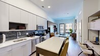 This well-appointed single-floor residence showcases a thoughtfully designed open-concept layout that seamlessly connects the kitchen, dining, and living areas. The kitchen is outfitted with sleek flat-front cabinetry in a crisp white finish, a full suite of stainless steel appliances including a side-by-side refrigerator, range, microwave, and dishwasher, and a striking blue-gray subway tile backsplash. A freestanding island with a white countertop provides additional workspace and casual dining space. Light wood-tone flooring flows throughout, unifying the living spaces with a warm, contemporary feel. The living area features an accent wall with warm wood slat paneling, a built-in open shelving unit, and a playful geometric wallpaper detail, creating a stylish focal point. Floor-to-ceiling glass doors open directly onto a private balcony, flooding the space with natural light and offering open views of the surrounding urban environment. The bedroom continues the balcony access theme, with large windows and a glass door that invite natural light and outdoor views. A dedicated walk-in closet with wire shelving and ample hanging space connects conveniently to the full bathroom, which features a soaking tub with a tiled surround, a dark wood vanity with a white countertop, and light porcelain tile flooring. Recessed lighting throughout enhances the clean, modern ambiance of the entire residence.