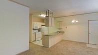 This single-story space showcases an open-concept layout that integrates the kitchen and living area into a cohesive, functional environment. The kitchen is equipped with light wood cabinetry that lines the walls, providing ample storage alongside a white electric range, microwave, and refrigerator. A central peninsula with a dark countertop extends into the space, offering additional workspace and a natural division between the kitchen and the carpeted living area. An overhead cabinet structure suspended above the peninsula adds a distinctive architectural element, enhancing vertical interest in the room. A sink is positioned along the peninsula, making the workspace both practical and accessible. Adjacent upper cabinetry on the far wall provides further storage. The living area features neutral wall-to-wall carpeting that contributes to a warm, comfortable atmosphere. A decorative pendant light fixture hangs near the living space, adding a subtle design accent. The overall color palette is light and neutral throughout, with cream-toned walls and cabinetry creating a bright, airy feel. The space transitions smoothly from the tiled kitchen floor to the carpeted living zone, clearly delineating each functional area while maintaining an open, connected feel.