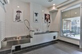 Located in Atlanta, GA, this single-floor pet spa is a thoughtfully designed grooming facility built with both functionality and comfort in mind. The space is fully enclosed with floor-to-ceiling glass walls and aluminum-framed doors, offering transparency between the interior grooming area and an adjacent outdoor deck, which is visible through large windows and features seating for pet owners.

The interior is finished with dark ceramic floor tiles and crisp white subway tile walls, creating a clean, clinical aesthetic well-suited to a grooming environment. A built-in stainless steel grooming tub is the centerpiece of the space, elevated on a tiled platform with convenient step access for pets. A professional-grade flexible spray hose with faucet and grooming supplies are mounted directly at the station, ensuring ease of use.

A wall-mounted split air conditioning unit provides climate control, keeping the environment comfortable year-round. Recessed ceiling lighting ensures the space is well-lit throughout. Framed artwork featuring dog photography adds a welcoming touch to the otherwise utilitarian design. A posted rules and guidelines sign near the entrance reinforces the space's shared, amenity-style function.