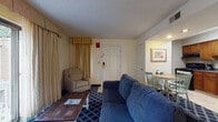 Located at 246 Daniel Webster Highway in Merrimack, New Hampshire, this hotel suite offers a practical and comfortable layout designed for both short and extended stays. The open-concept living and sleeping area is anchored by a king-sized bed dressed in crisp white linens, flanked by matching nightstands with warm lighting. A large flat-screen television sits atop a dark dresser, while a blue sofa and armchair create a welcoming lounge area, all unified by a bold blue diamond-patterned carpet. A wooden coffee table completes the sitting arrangement. The full kitchen is equipped with warm wood cabinetry, a full-sized refrigerator, dishwasher, stovetop range, microwave, and ample counter space — providing everything needed for in-suite meal preparation. A small dining table with chairs sits adjacent to the kitchen, offering a casual spot for meals. The bathroom features a wide vanity with dual mirrors, a well-lit vanity bar, and wood cabinetry providing generous storage. Drapes frame the windows throughout, allowing for privacy and light control. The suite's single-floor layout ensures easy accessibility, while its cohesive design and comprehensive amenities make it a fully functional and comfortable accommodation in Merrimack, NH.