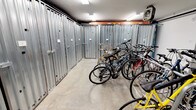 This dedicated bike room offers a practical and well-organized space designed to accommodate the storage needs of cyclists. The room is lined along its walls with rows of galvanized steel corrugated storage lockers, each fitted with individual doors and locks, providing secure, private storage for bicycles or other personal equipment. Open bicycle racks run through the center and along the sides of the room, offering additional space for easy-access bike parking and accommodating a variety of bicycle styles, from road bikes to mountain bikes. The concrete flooring is durable and easy to maintain, well-suited to the utilitarian nature of the space. Overhead fluorescent lighting ensures the room is brightly and evenly illuminated throughout. Exposed ceiling infrastructure, including piping and structural beams, reflects the functional, no-frills design of the space. A wall-mounted heater is present, helping to maintain a stable environment year-round. Access doors are positioned conveniently within the room, and a fire extinguisher is mounted on the wall, underscoring the attention to safety. Overall, this bike room delivers a secure, organized, and accessible solution for bicycle storage.