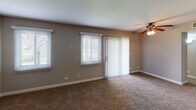 Located at 214 Washington Square in Elk Grove Village, Illinois, this single-floor residence offers a well-organized and functional layout throughout. The living area is generously sized, featuring warm neutral-toned walls, plush carpeting, and a ceiling fan with light fixture. Natural light fills the space through a combination of horizontal blinds on double windows and a sliding glass door dressed with vertical blinds, providing easy access to the outdoors. The open flow connects the living area to the kitchen, which is thoughtfully appointed with warm oak cabinetry, granite-style countertops, a stainless steel refrigerator, and a stainless steel sink. A mosaic tile backsplash in shades of gray and brown adds a decorative touch, complemented by hardwood-style flooring that grounds the kitchen space. The bedroom is a comfortable, carpeted retreat with a window fitted with horizontal blinds that allow for natural light and outdoor views. The full bathroom features tile flooring, a wood-toned vanity with light countertop, a standard tub-shower combination, and neutral wall tones that carry throughout the home. Clean white trim and baseboards provide crisp contrast against the taupe walls, lending the overall space a cohesive and move-in ready appearance.