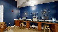 Located at 8800 Pine Forest Rd in Pensacola, Florida, this single-floor community space is thoughtfully designed to serve as both a relaxing lounge and a functional business center. The interior is defined by striking navy blue accent walls adorned with decorative gold metalwork floral sculptures, creating a bold and sophisticated visual identity throughout.

The business center area features a long built-in workstation with warm wood cabinetry, dark granite countertops, multiple computer terminals, and a multifunction printer — all illuminated by an elegant drum pendant light. Gold metal bar stools complement the cabinetry's rich tones.

A cozy reading nook offers two tufted leather armchairs arranged on a vintage-style area rug, flanked by a well-stocked bookshelf and a tall arc floor lamp. Large windows with white plantation shutters flood this corner with natural light, enhancing the inviting atmosphere.

The open-plan layout transitions seamlessly into a broader common area with light wood flooring, recessed lighting, and stately dark wood square columns that add architectural character. Tall ceilings amplify the sense of space, while additional windows bring in ample natural light. Decorative vases and thoughtful furnishings throughout contribute to the polished, welcoming ambiance of this well-appointed community gathering space.