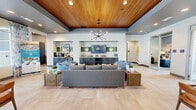 Located in Dacula, Georgia, this single-floor community clubhouse offers a thoughtfully designed space that balances functionality with contemporary style. The layout encompasses a central living area, multiple hallways, and dedicated office spaces, all unified by a cohesive modern aesthetic.

The main living area serves as the heart of the space, featuring warm wood-paneled ceilings that contrast beautifully with the light large-format tile flooring. A statement sputnik chandelier anchors the room, while built-in cabinetry with green tile accents and a mounted display screen create an inviting focal point. Plush gray sectional seating and accent chairs are arranged to encourage conversation and comfort.

The office area is distinguished by floor-to-ceiling glass windows and glazed double doors that flood the space with natural light and provide sweeping views of the outdoor courtyard. A dark-toned ceiling with a bold pendant light adds a sophisticated contrast to the airy, light-filled atmosphere, while floral drapery panels add a touch of softness.

The connecting hallways are clean and efficiently designed, featuring light neutral walls, tile flooring, and glass-paneled office entry doors with clear signage. The overall ambiance throughout is bright, polished, and welcoming — reflecting a well-appointed community facility designed for both professional and social use.