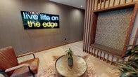 This contemporary lobby makes an immediate impression with its thoughtfully curated design and bold personality. At the heart of the space, a striking neon sign reading "live life on the edge" commands attention against a taupe textured wall, setting an energetic and aspirational tone for the entire environment.

A custom wood slat partition serves as a defining architectural feature, framing a built-in upholstered nook accented with an ornate patterned wallcovering. This warm, natural wood element provides a visual anchor and subtly divides the space while maintaining an open and airy feel. Light travertine-style tile flooring runs throughout, complementing the neutral wall tones and warm wood accents.

The seating area near the entrance features a pair of rich cognac leather chairs positioned to take full advantage of the floor-to-ceiling glass storefront, which floods the lobby with natural light and offers views of mature trees and the surrounding streetscape. A hammered brass side table atop a cowhide rug adds a layer of texture and artisanal character.

An overhead sputnik-style chandelier provides ambient illumination, while recessed lighting ensures the space is evenly and warmly lit throughout. A sculptural art piece displayed on a pedestal near the entry and lush indoor greenery further enrich the space. A long corridor visible from the lobby suggests additional amenities beyond.