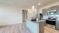 This single-floor residence is designed with a clean, contemporary aesthetic and functional layout. The kitchen serves as the centerpiece of the living area, anchored by a large granite-topped island with an integrated sink and elegant pendant lighting overhead. Gray shaker-style cabinetry lines the walls, complemented by a white subway tile backsplash and a full suite of stainless steel appliances, including a refrigerator, range, and microwave. Light wood-style flooring runs throughout the main living areas, enhancing the bright, open feel of the space.

The bedrooms feature soft carpeting and are bathed in natural light through large windows fitted with blinds. Ceiling fans provide additional comfort, and the neutral white walls offer a versatile backdrop. The bathroom is finished with large-format marble-style wall tiles surrounding a soaking tub and shower combination, paired with light wood-look flooring, a granite vanity top, and gray cabinetry that coordinates with the kitchen's design language.

A dedicated laundry area accommodates a full-size top-loading washer and includes a built-in shelf above for added storage. Throughout the unit, consistent use of neutral tones, modern fixtures, and quality finishes creates a cohesive and comfortable living environment.