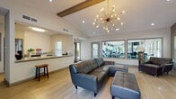 This well-appointed single-floor community clubhouse offers a thoughtfully designed gathering space that blends comfort, function, and modern aesthetics. The main lounge area serves as the heart of the space, anchored by a tufted leather sectional sofa and accent chairs arranged around a tile-surround fireplace with a wall-mounted television above. Soaring vaulted ceilings with exposed wood beams add architectural character, while a striking sputnik chandelier provides a bold focal point and warm ambient lighting. Light-toned hardwood-style flooring runs throughout, creating a clean and cohesive look. Large windows fitted with horizontal blinds allow ample natural light to filter in while offering views of surrounding greenery. Adjacent to the lounge, a kitchenette with granite-style countertops and a sink offers convenient refreshment access. A dedicated workspace or meeting nook features a modern table and chairs set beside multiple windows, making it ideal for focused work or small gatherings. Interior windows between the lounge and the adjoining fitness center provide a visual connection to the well-equipped gym, which is visible with cardio machines and workout equipment. The overall ambiance is contemporary and inviting, designed to serve as a versatile shared amenity space.