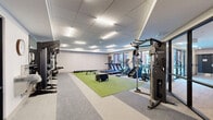 Located at 100 Mia Rose Way in Dardenne Prairie, Missouri, this well-appointed fitness center offers a thoughtfully designed space for a full range of exercise activities. The single-floor layout makes efficient use of its generous footprint, organizing equipment into distinct functional zones that flow naturally throughout the room.

The space features a drop-panel ceiling with recessed linear lighting that provides bright, even illumination across the entire floor. Large glass panel walls and windows along one side allow natural light to filter in while offering views to adjacent common areas and outdoor spaces, creating an open, connected atmosphere.

The flooring combines neutral carpet tiles with a vibrant turf training surface at the center of the room, which serves as the focal point for functional fitness and athletic training activities. Plyo boxes of varying heights are positioned on the turf alongside a flat bench and kettlebells, supporting dynamic training routines.

Along the perimeter, the space is equipped with a full dumbbell rack, medicine balls, a multi-station cable machine by TKO, and a power rack with weight plates. The cardio section features multiple treadmills, stair climbers, and elliptical machines, all positioned near the windows to maximize natural light during workouts. A water fountain station is conveniently mounted on the wall near the glass entry doors, completing this comprehensive and modern fitness facility.