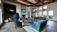 This single-floor amenity lounge is a well-appointed gathering space that seamlessly blends comfort and sophistication. The room's most striking architectural feature is its soaring coffered ceiling adorned with rich, dark wood beams arranged in a geometric grid pattern, lending a warm and refined character to the space. Large windows along the perimeter flood the interior with natural light, creating an inviting and open atmosphere.

The lounge is thoughtfully divided into distinct zones for leisure and socializing. A central seating area anchors the space, featuring a mix of bold navy wingback chairs, tufted lavender accent chairs, and a teal sofa arranged around a minimalist wood-and-metal coffee table, accented by tasteful greenery. A wall-mounted flat-screen television is positioned above a tiled fireplace, which serves as a warm, eye-catching focal point. Adjacent to the seating area, a full-size billiards table provides an additional entertainment option, set beneath elegant pendant lighting. Decorative sculptural elements and layered rugs over slate-style tile flooring add texture and visual depth throughout. The overall ambiance is one of upscale comfort, designed to encourage both casual relaxation and social engagement.