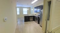 This single-floor space showcases a thoughtfully designed open-concept layout that maximizes both functionality and visual appeal. The kitchen serves as the centerpiece, featuring sleek dark cabinetry that contrasts elegantly with light granite countertops. A suite of stainless steel appliances — including a refrigerator, electric range, built-in microwave, and dishwasher — provides a fully equipped cooking environment. Pendant lights hang above the kitchen area, adding a modern decorative touch while providing focused task lighting. The light-toned hardwood-style flooring runs seamlessly throughout, contributing to a clean, cohesive aesthetic. Soft sage-green walls lend a calm, neutral ambiance to the interior. Natural light filters in through multiple windows fitted with horizontal blinds, brightening the open space without sacrificing privacy. The kitchen opens naturally into the adjoining living area, creating an inviting flow that makes the space feel open and airy. A hallway visible from the main area suggests well-organized access to additional rooms. Overall, the space combines practical design with contemporary finishes, offering a comfortable and modern environment well-suited for everyday living.