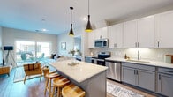 Located at 6570 Hearthstone Ct NW in Rochester, MN, the Telluride floor plan offers a well-designed single-floor layout with 2 bedrooms, 2 bathrooms, and 2 closets. The open-concept kitchen is a standout feature, showcasing two-tone cabinetry with white upper and gray lower cabinets, quartz countertops, stainless steel appliances, and a central island with seating — all illuminated by striking black pendant lights. The kitchen flows seamlessly into the living room, which is bright and airy thanks to large sliding glass doors that open to a private patio with views of the surrounding neighborhood. The primary bedroom is a comfortable retreat with plush carpeting, a large window for natural light, and direct access to a well-appointed en suite bathroom featuring dual sinks, quartz countertops, gray cabinetry with matte black hardware, and a walk-in closet with built-in wire shelving. The second bedroom is equally comfortable, with soft carpeting and access to a full bathroom complete with a tub/shower combo, gray vanity, and wood-look luxury vinyl flooring. Throughout the home, light-toned walls and consistent modern finishes create a clean, cohesive aesthetic.