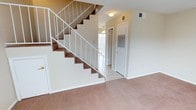This two-story residence offers a straightforward and functional layout across both levels. The main floor greets visitors with a spacious carpeted living area that provides a warm, comfortable feel underfoot. A centrally positioned staircase with clean white metal railings and carpeted treads serves as both a practical and visual centerpiece, connecting the two floors with ease.

The main level transitions from carpet to tile flooring near the entryway and hallway, offering a clear delineation between living and transitional spaces. A ceiling fan is visible toward the rear of the lower level, promoting airflow throughout the common areas. Multiple HVAC vents are positioned throughout the space, reflecting a well-considered climate control system.

Natural light filters in through a window or glass door at the rear of the main floor, brightening the entry corridor and stairwell area. The neutral color palette — soft beige walls, white trim, and warm-toned carpet — creates a clean and versatile backdrop suitable for a variety of furnishing styles.

Overall, the space presents a practical and livable design with consistent finishes across both floors, making it well-suited for everyday residential use.