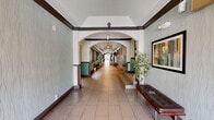 This single-floor corridor space spans a generous footprint, designed to serve as a functional and visually appealing communal hallway. The layout is long and linear, connecting multiple sections through a series of elegant arched passageways that add architectural character and a sense of depth throughout the space.

The flooring consists of large-format, light-toned tiles laid uniformly across the entire length of the corridor, contributing to a clean and polished appearance. The walls are finished with a textured, muted sage-green wallcovering accented by dark wood crown molding and baseboards, creating a refined contrast that runs consistently throughout.

Ceiling fans are installed at regular intervals along the corridor, providing both air circulation and ambient lighting. The ceilings feature tray-style designs that add subtle architectural dimension. Windows positioned along one side of the hallway allow natural light to filter in, offering views of exterior greenery.

Practical amenities are thoughtfully integrated, including multiple banks of individual metal mailboxes and parcel lockers installed along the walls. Potted plants and decorative artwork are placed throughout, softening the utilitarian elements and enhancing the overall ambiance. A cushioned bench provides a seating area, rounding out this well-maintained and functional communal space.